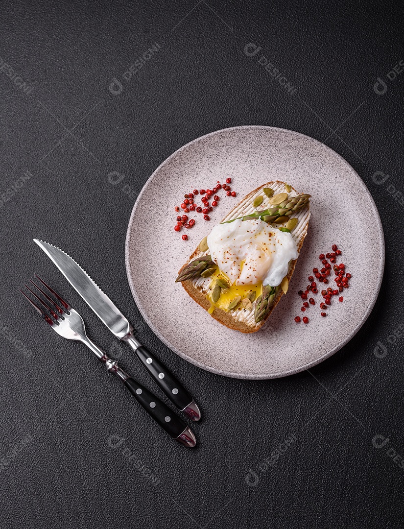 Delicioso café da manhã composto por ovos escalfados com torradas com cream cheese, aspargos, sal e especiarias em um fundo escuro de concreto