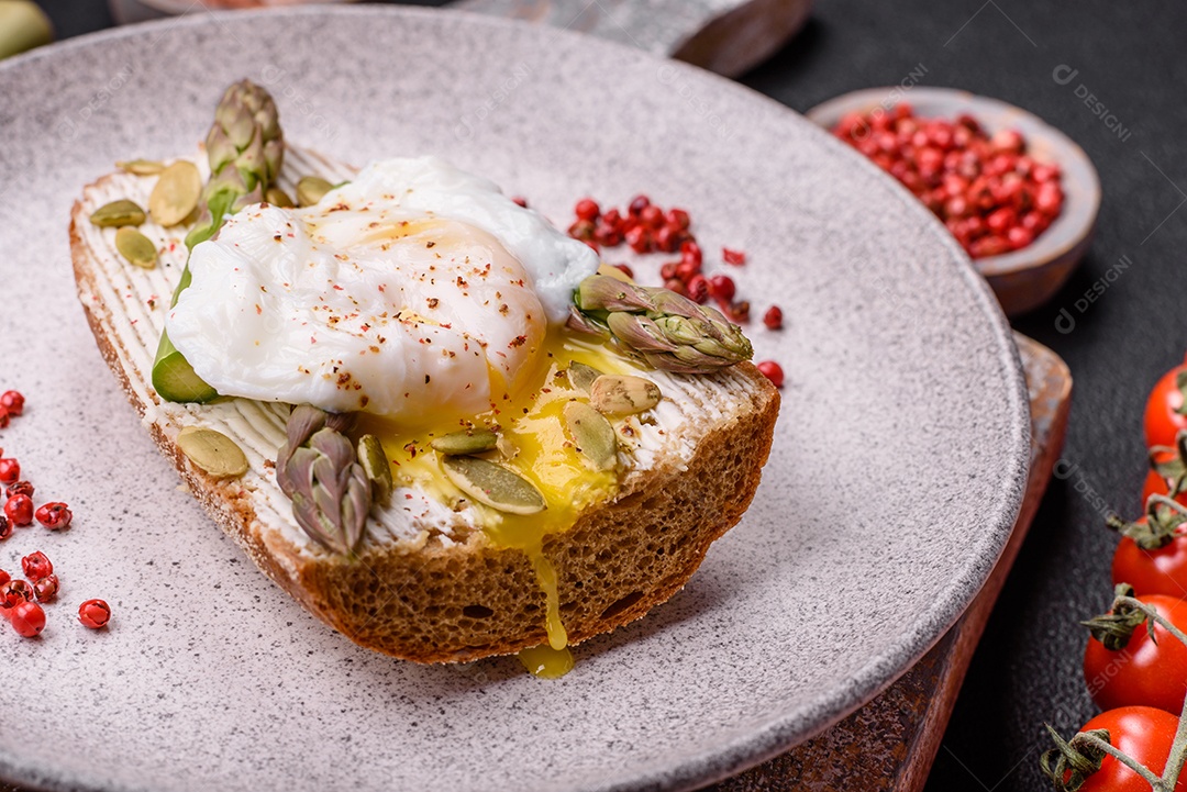 Delicioso café da manhã composto por ovos escalfados com torradas com cream cheese, aspargos, sal e especiarias em um fundo escuro de concreto