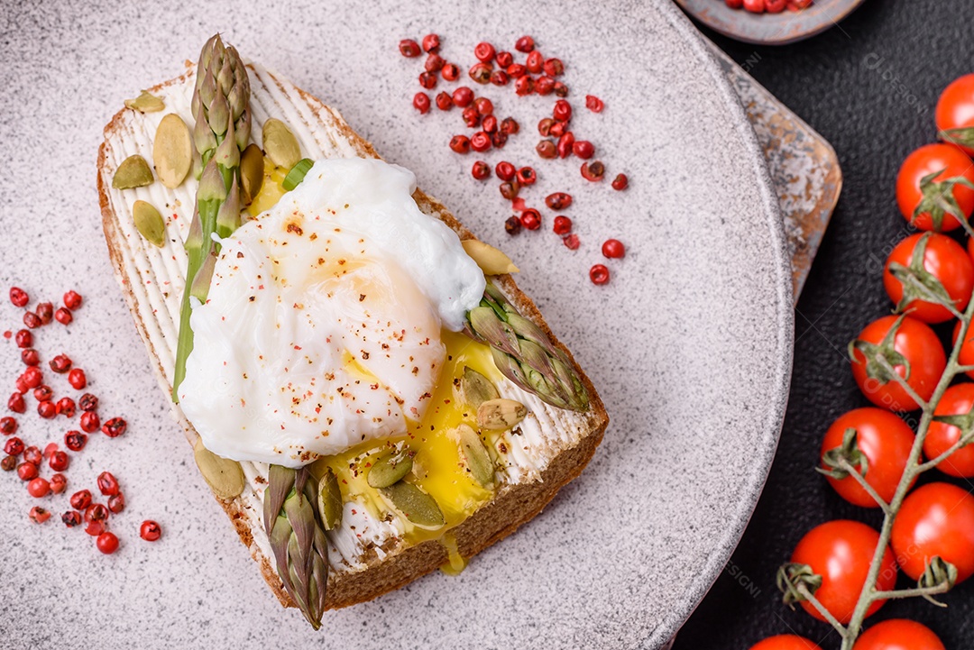 Delicioso café da manhã composto por ovos escalfados com torradas com cream cheese, aspargos, sal e especiarias em um fundo escuro de concreto