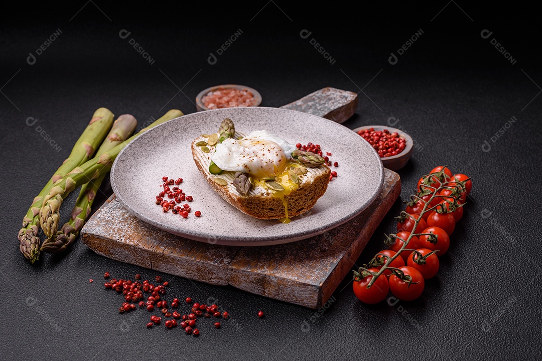 Delicioso café da manhã composto por ovos escalfados com torradas com cream cheese, aspargos, sal e especiarias em um fundo escuro de concreto