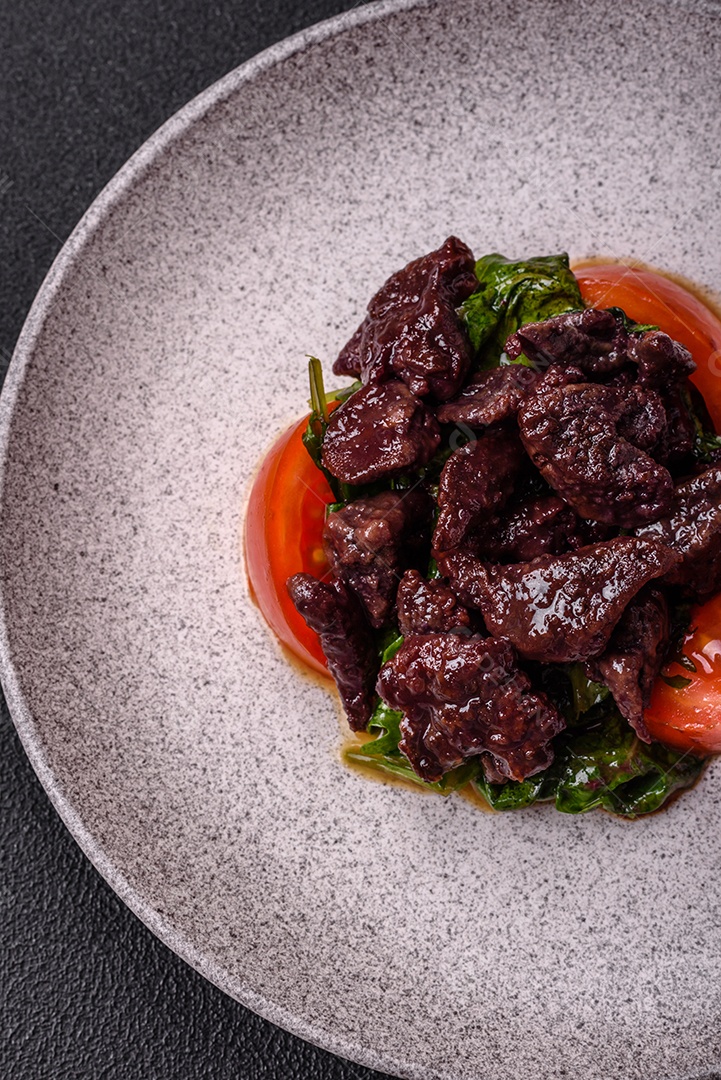 Deliciosa salada quente de fígado de galinha, espinafre, rúcula, tomate com sal, especiarias e ervas sobre um fundo escuro de concreto