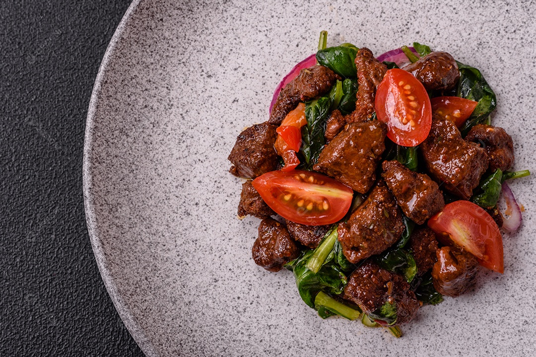 Deliciosa salada quente com pedaços de carne, espinafre, tomate, sal, especiarias e ervas sobre um fundo escuro de concreto