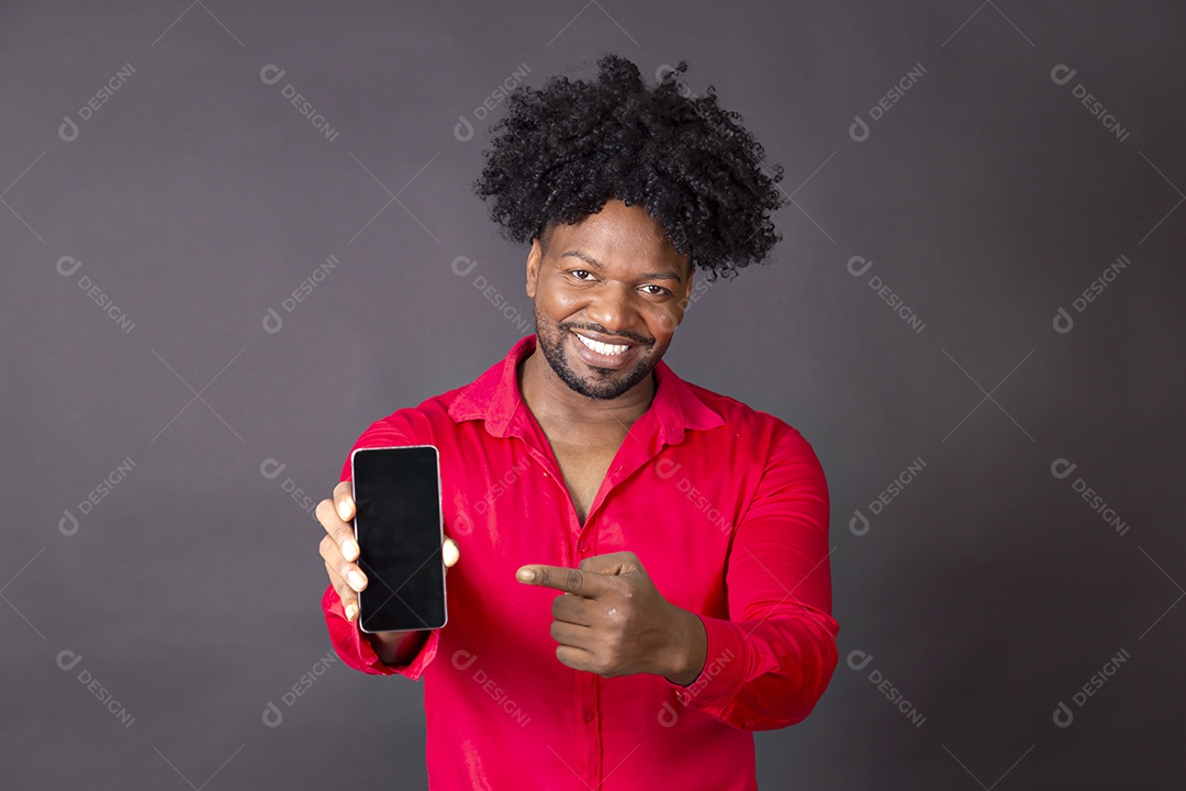 Um homem moreno segurando celular e sorrindo apontando para ele