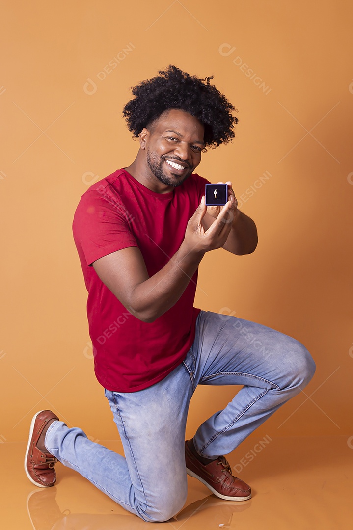Um homem moreno de camiseta vermelha ajoelhado com caixa de aliança