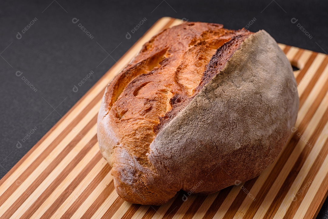 Um pedaço de pão integral com grãos de cereais em uma tábua de madeira sobre um fundo escuro de concreto