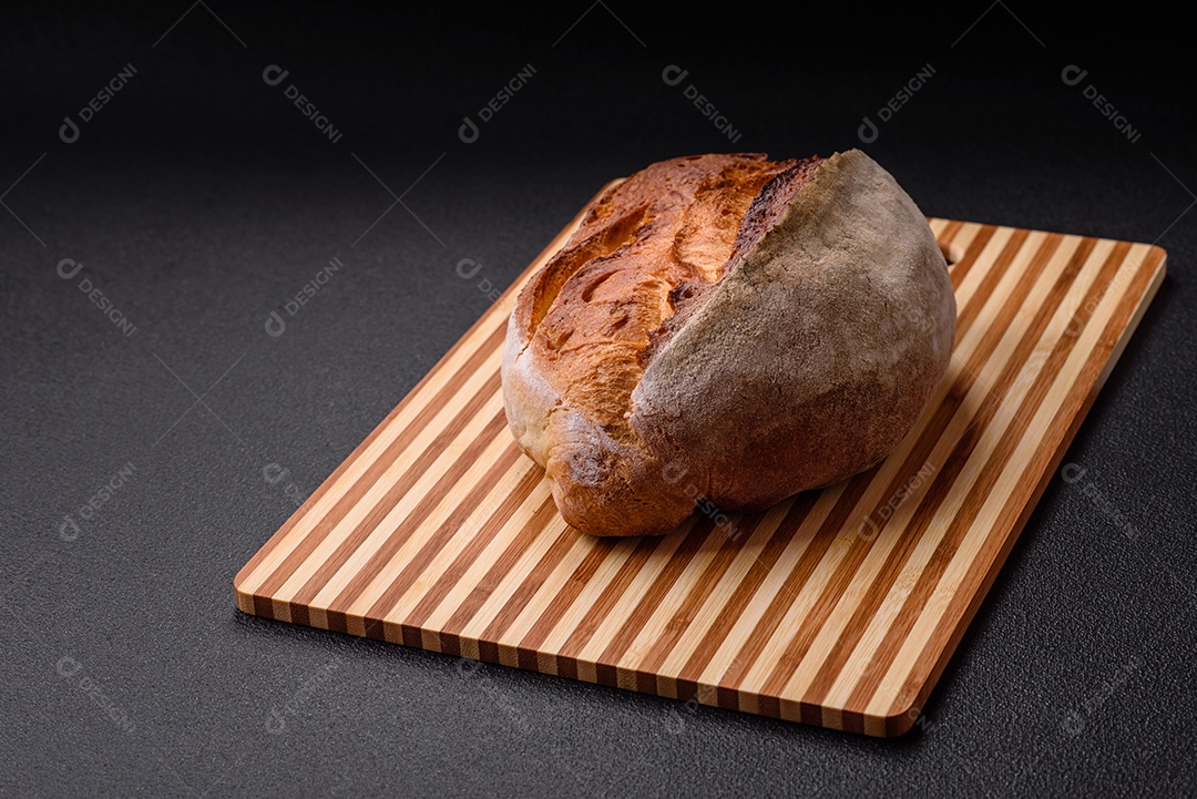 Um pedaço de pão integral com grãos de cereais em uma tábua de madeira sobre um fundo escuro de concreto