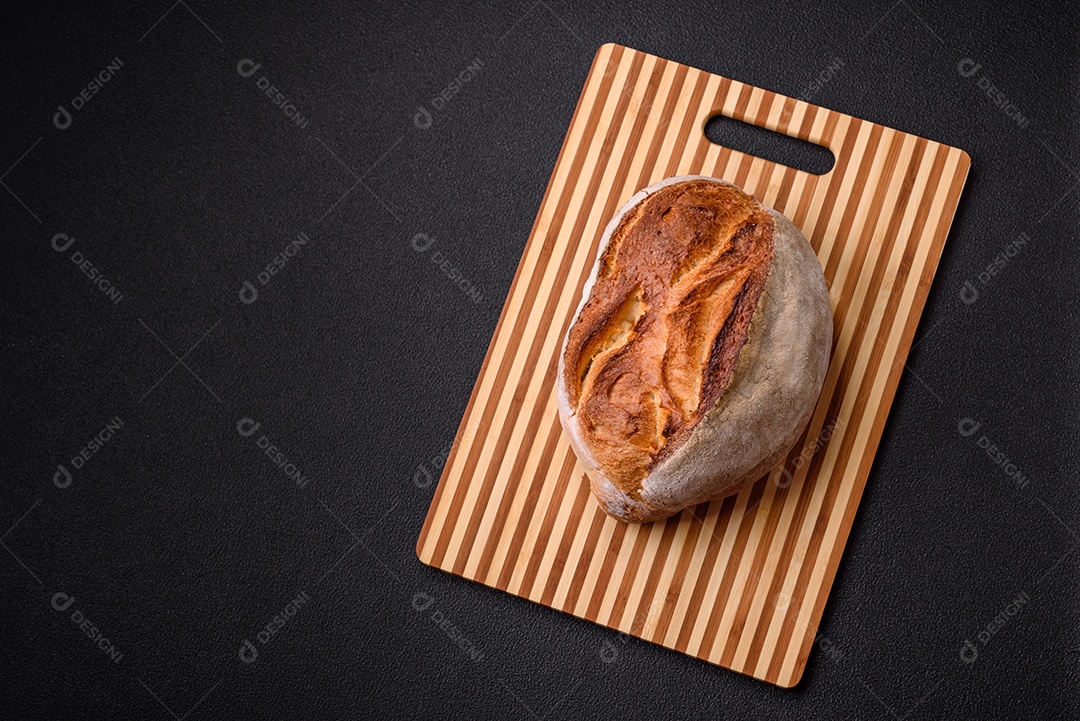 Um pedaço de pão integral com grãos de cereais em uma tábua de madeira sobre um fundo escuro de concreto