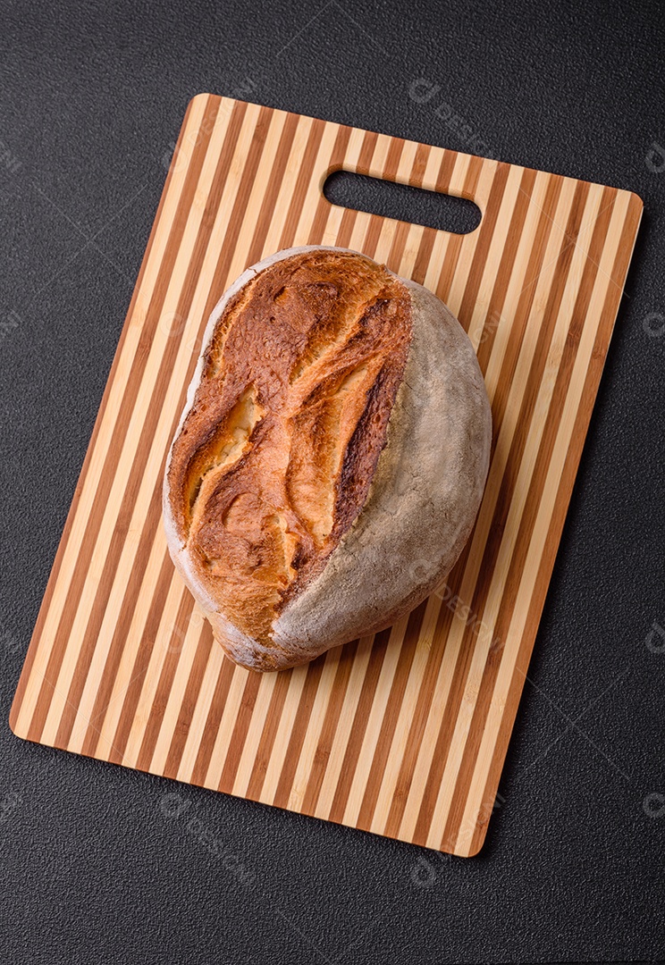 Um pedaço de pão integral com grãos de cereais em uma tábua de madeira sobre um fundo escuro de concreto
