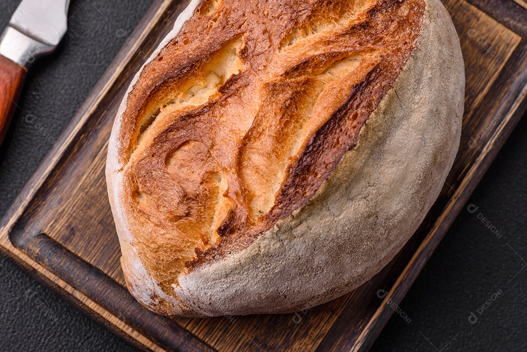 Um pedaço de pão integral com grãos de cereais em uma tábua de madeira sobre um fundo escuro de concreto