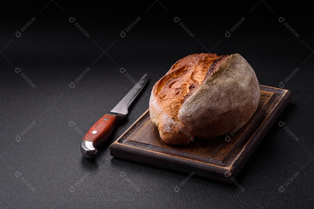 Um pedaço de pão integral com grãos de cereais em uma tábua de madeira sobre um fundo escuro de concreto