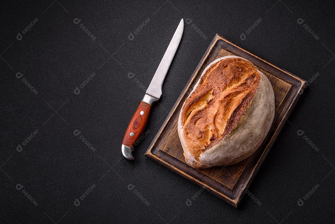 Um pedaço de pão integral com grãos de cereais em uma tábua de madeira sobre um fundo escuro de concreto