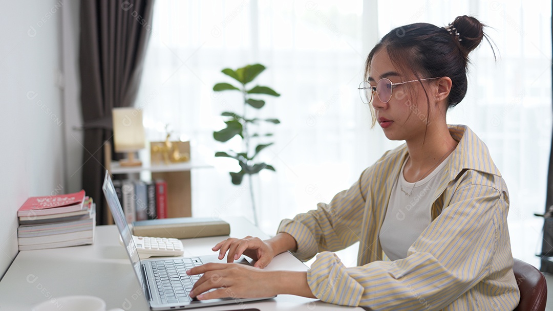 Jovem está sentada em uma mesa digitando trabalho em seu laptop