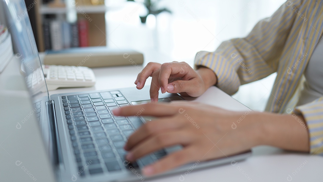 Uma Jovem asiática está sentada em sua mesa concentrada na tela e digitando no laptop