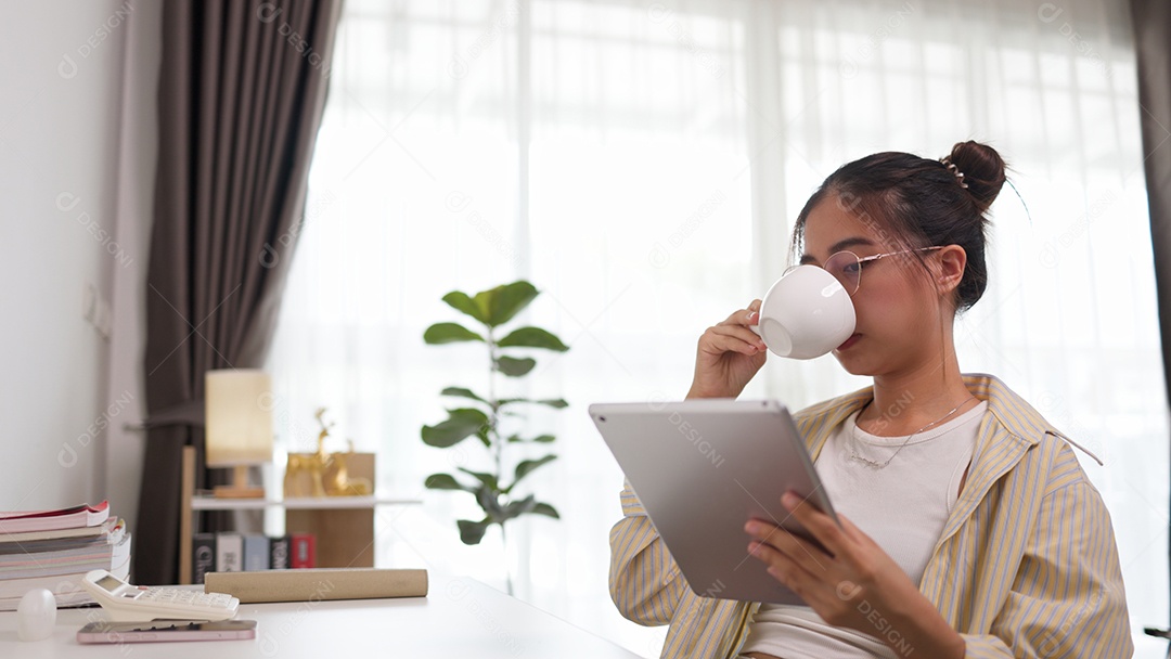 Mulher asiática usando seu laptop pega uma xícara de café e toma um gole