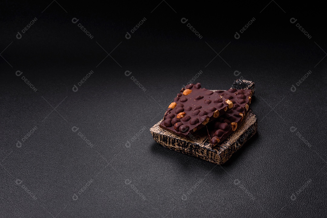 Chocolate amargo preto com avelãs inteiras, frutas cristalizadas e hortelã, quebrado em pedaços sobre fundo escuro de concreto