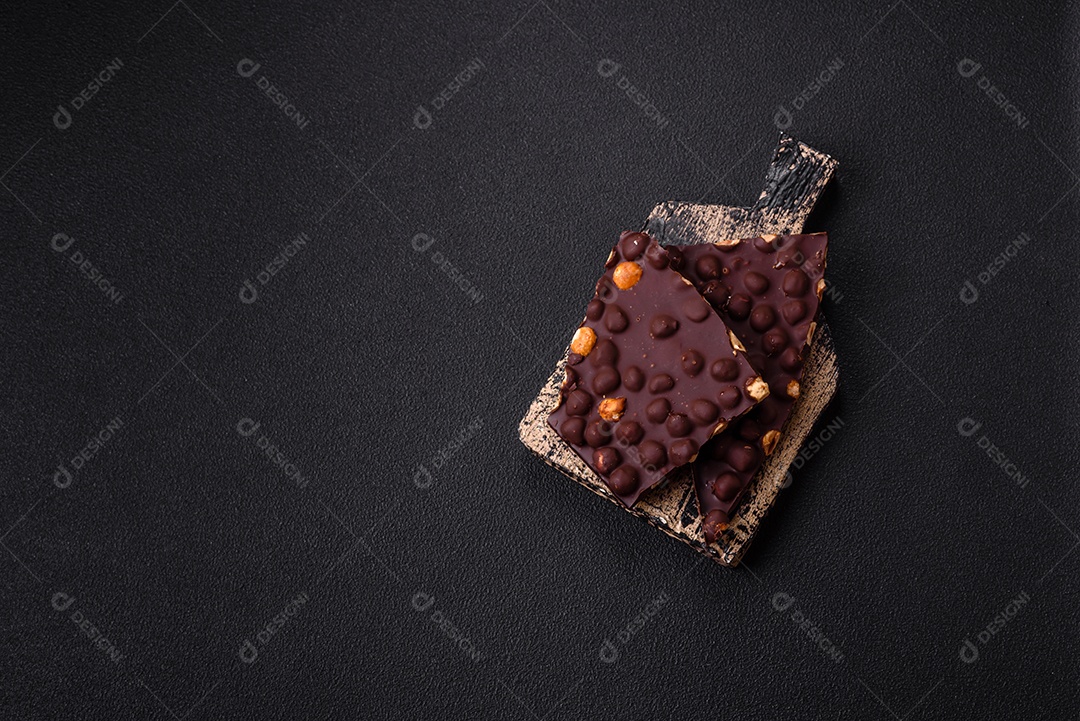 Chocolate amargo preto com avelãs inteiras, frutas cristalizadas e hortelã, quebrado em pedaços sobre fundo escuro de concreto
