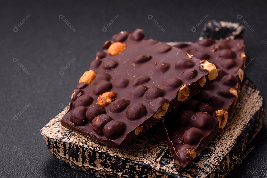 Chocolate amargo preto com avelãs inteiras, frutas cristalizadas e hortelã, quebrado em pedaços sobre fundo escuro de concreto