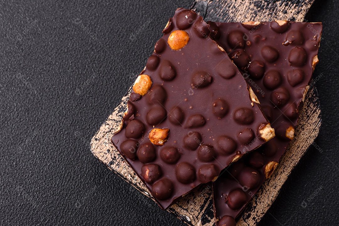 Chocolate amargo preto com avelãs inteiras, frutas cristalizadas e hortelã, quebrado em pedaços sobre fundo escuro de concreto