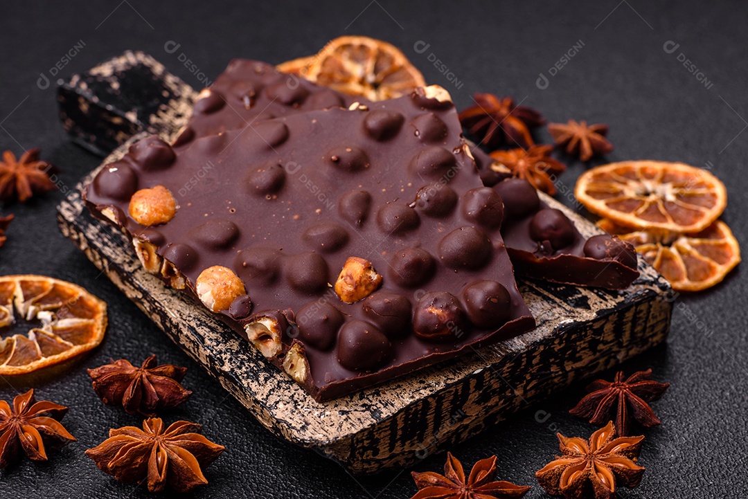 Chocolate amargo preto com avelãs inteiras, frutas cristalizadas e hortelã, quebrado em pedaços sobre fundo escuro de concreto