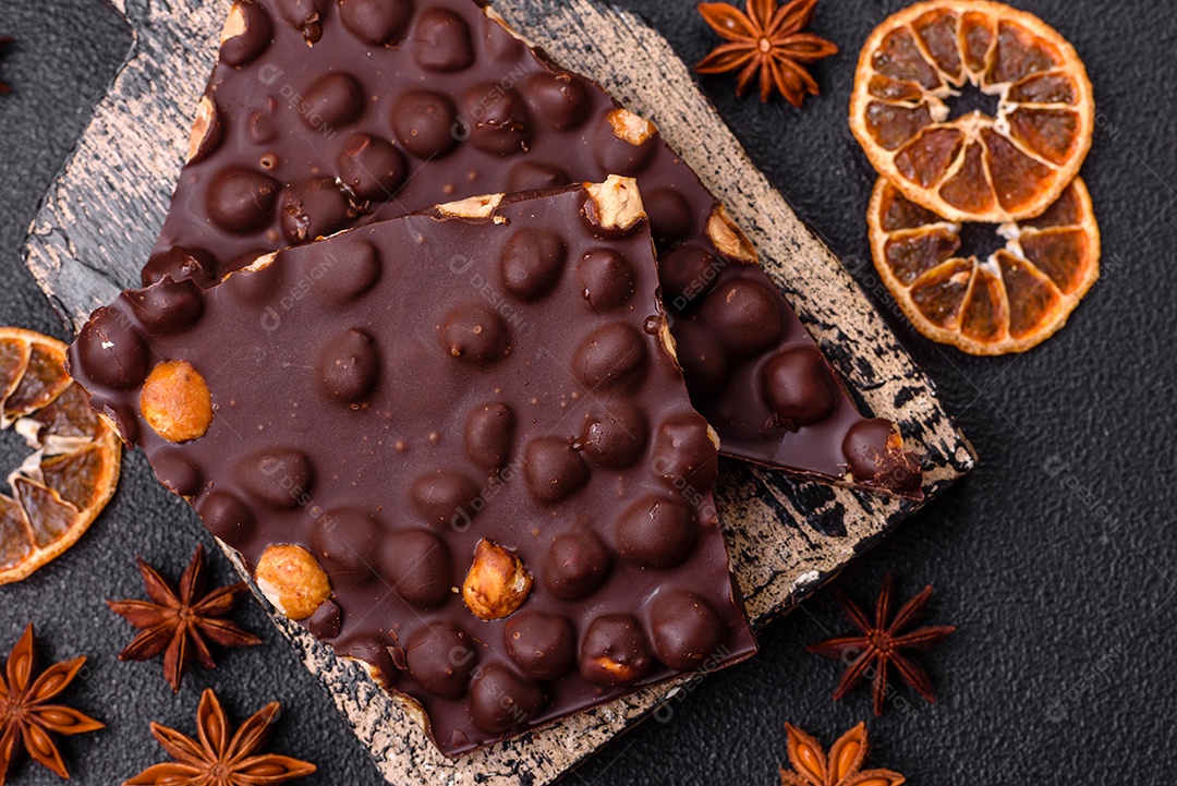Chocolate amargo preto com avelãs inteiras, frutas cristalizadas e hortelã, quebrado em pedaços sobre fundo escuro de concreto