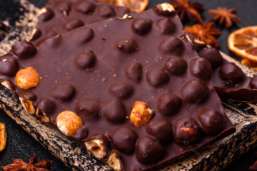 Chocolate amargo preto com avelãs inteiras, frutas cristalizadas e hortelã, quebrado em pedaços sobre fundo escuro de concreto