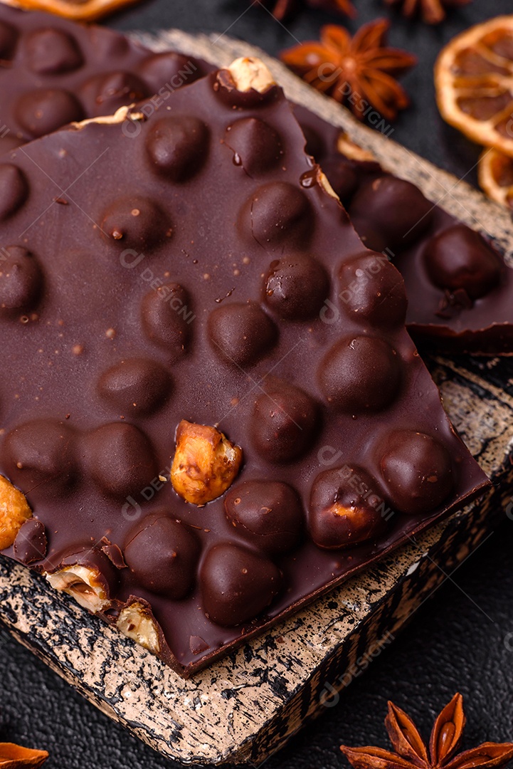 Chocolate amargo preto com avelãs inteiras, frutas cristalizadas e hortelã, quebrado em pedaços sobre fundo escuro de concreto