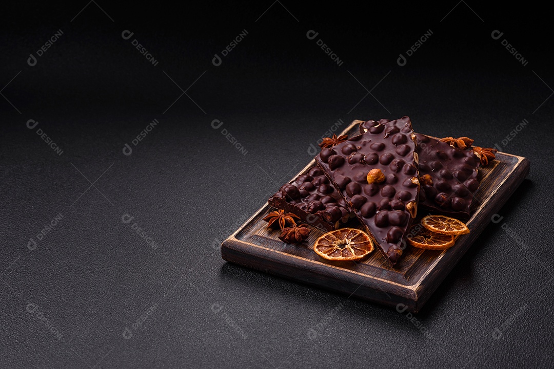 Chocolate amargo preto com avelãs inteiras, frutas cristalizadas e hortelã, quebrado em pedaços sobre fundo escuro de concreto