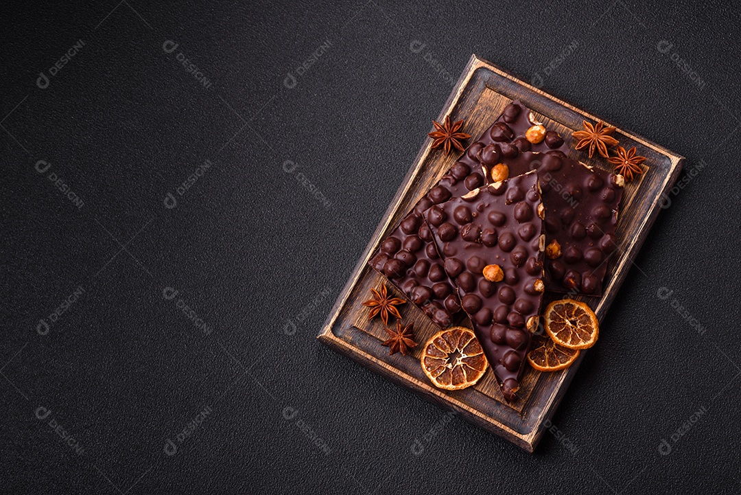 Chocolate amargo preto com avelãs inteiras, frutas cristalizadas e hortelã, quebrado em pedaços sobre fundo escuro de concreto
