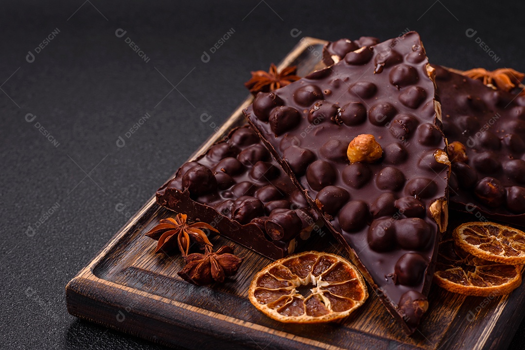 Chocolate amargo preto com avelãs inteiras, frutas cristalizadas e hortelã, quebrado em pedaços sobre fundo escuro de concreto