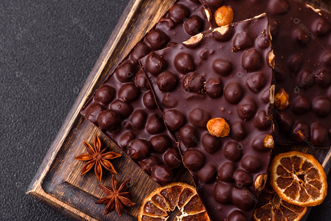 Chocolate amargo preto com avelãs inteiras, frutas cristalizadas e hortelã, quebrado em pedaços sobre fundo escuro de concreto