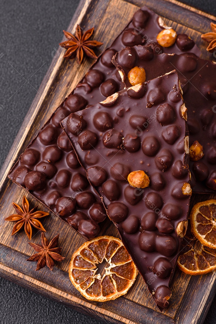 Chocolate amargo preto com avelãs inteiras, frutas cristalizadas e hortelã, quebrado em pedaços sobre fundo escuro de concreto