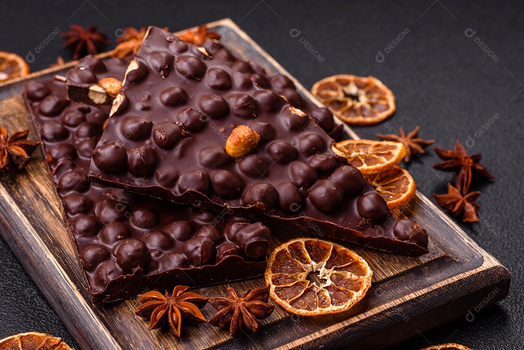 Chocolate amargo preto com avelãs inteiras, frutas cristalizadas e hortelã, quebrado em pedaços sobre fundo escuro de concreto