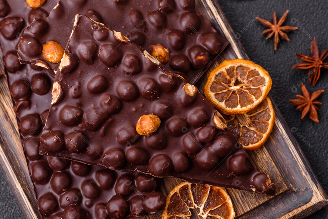 Chocolate amargo preto com avelãs inteiras, frutas cristalizadas e hortelã, quebrado em pedaços sobre fundo escuro de concreto