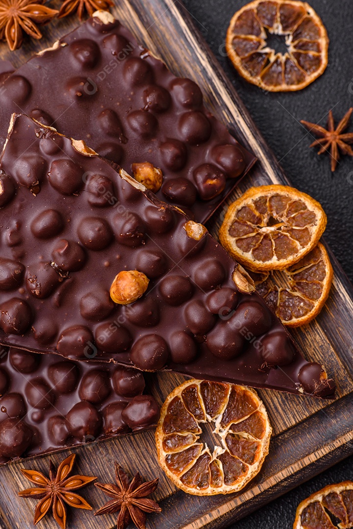 Chocolate amargo preto com avelãs inteiras, frutas cristalizadas e hortelã, quebrado em pedaços sobre fundo escuro de concreto