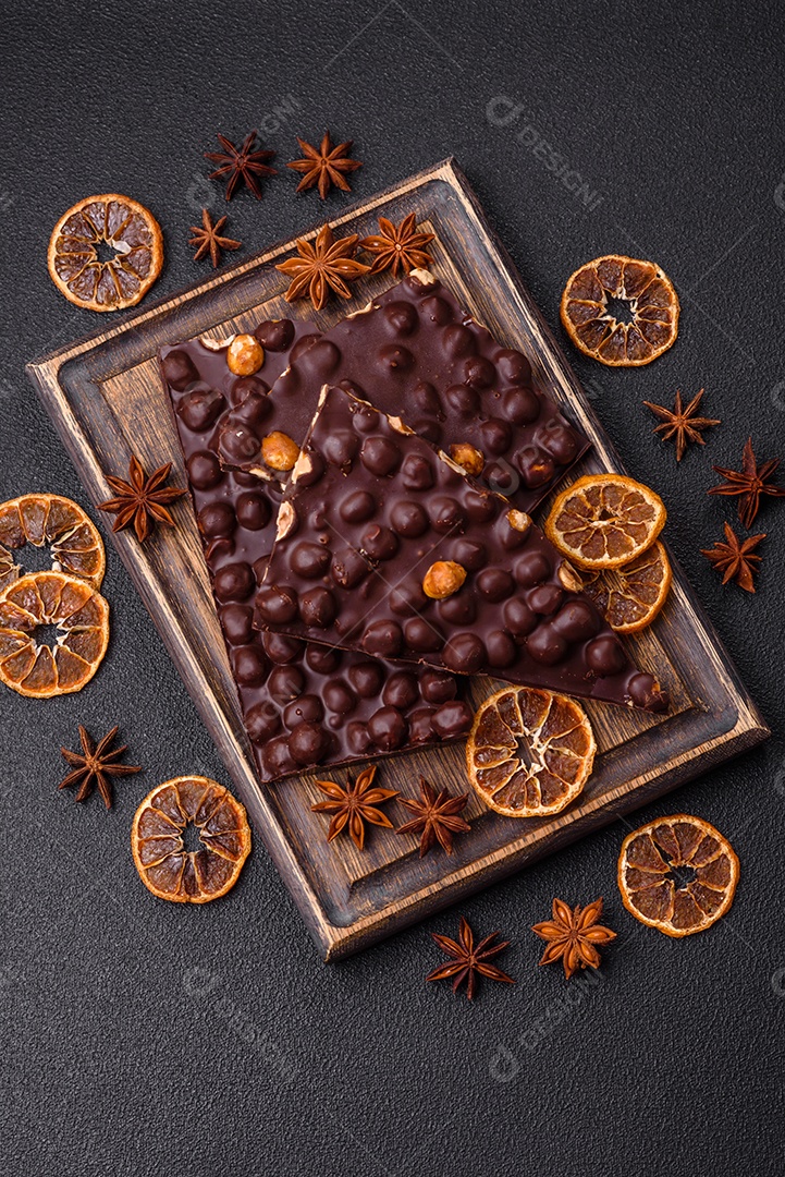 Chocolate amargo preto com avelãs inteiras, frutas cristalizadas e hortelã, quebrado em pedaços sobre fundo escuro de concreto