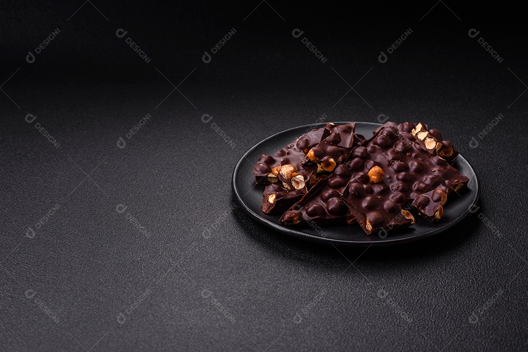 Chocolate amargo preto com avelãs inteiras, frutas cristalizadas e hortelã, quebrado em pedaços sobre fundo escuro de concreto