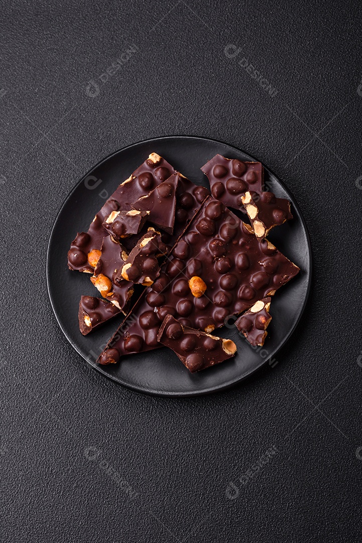 Chocolate amargo preto com avelãs inteiras, frutas cristalizadas e hortelã, quebrado em pedaços sobre fundo escuro de concreto