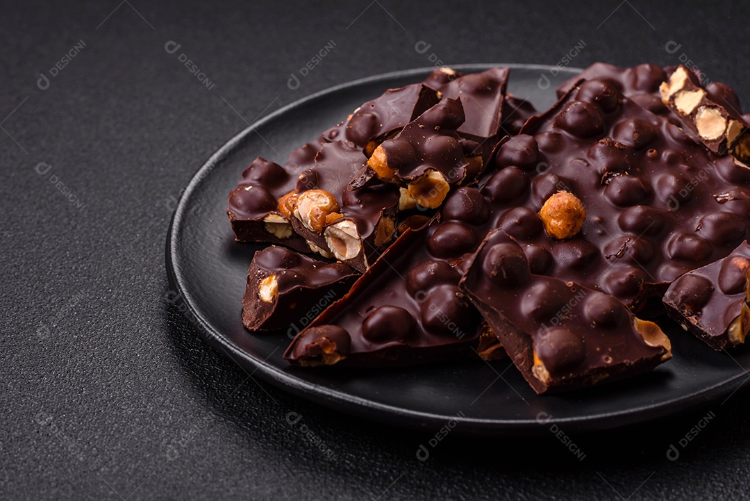 Chocolate amargo preto com avelãs inteiras, frutas cristalizadas e hortelã, quebrado em pedaços sobre fundo escuro de concreto