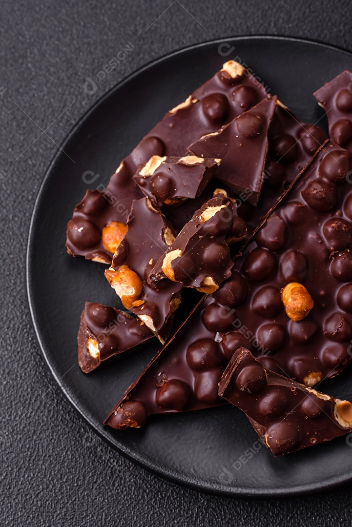 Chocolate amargo preto com avelãs inteiras, frutas cristalizadas e hortelã, quebrado em pedaços sobre fundo escuro de concreto