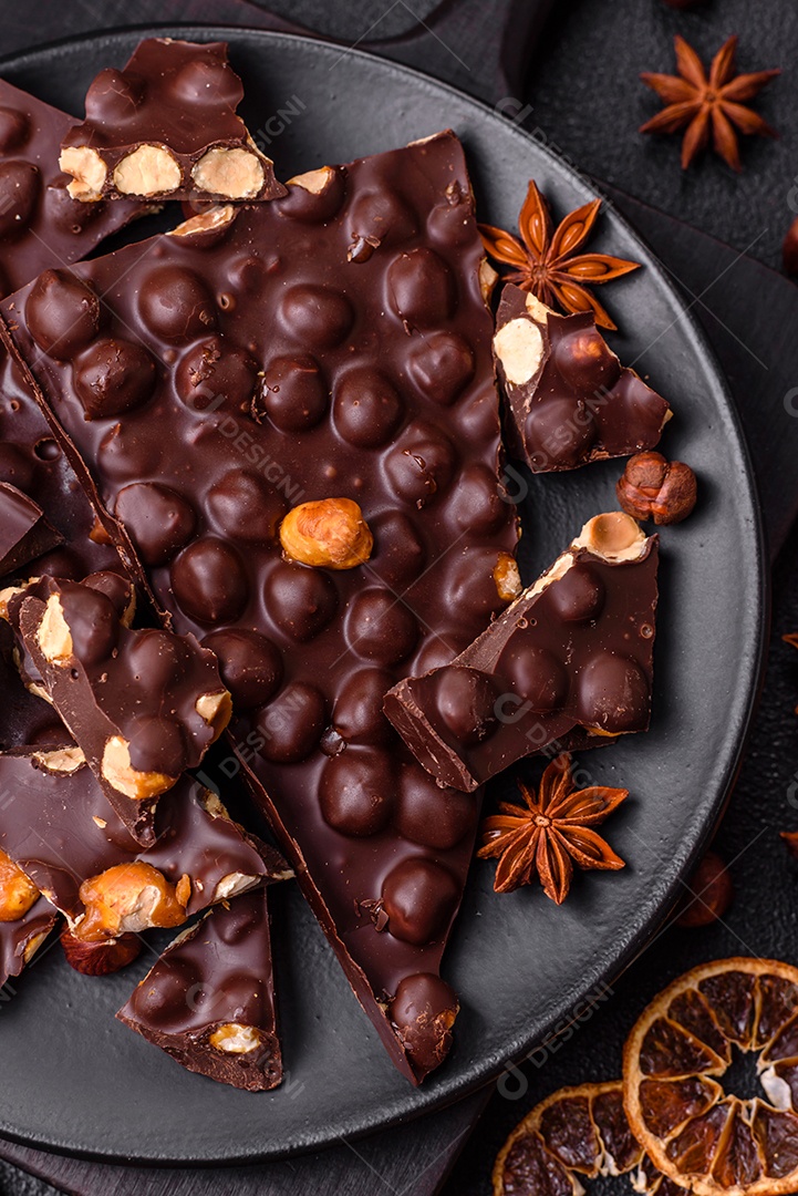Chocolate amargo preto com avelãs inteiras, frutas cristalizadas e hortelã, quebrado em pedaços sobre fundo escuro de concreto