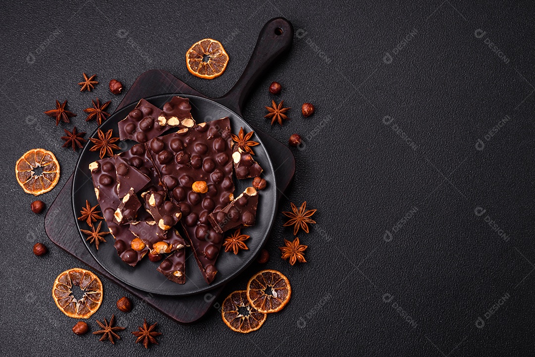 Chocolate amargo preto com avelãs inteiras, frutas cristalizadas e hortelã, quebrado em pedaços sobre fundo escuro de concreto