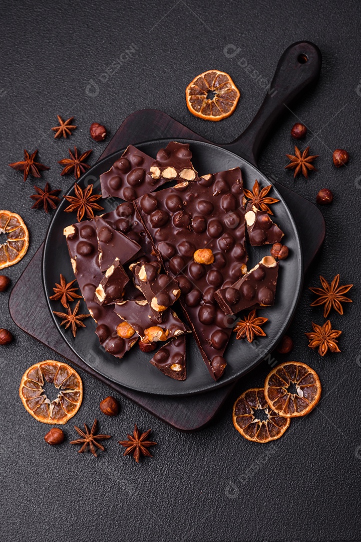Chocolate amargo preto com avelãs inteiras, frutas cristalizadas e hortelã, quebrado em pedaços sobre fundo escuro de concreto