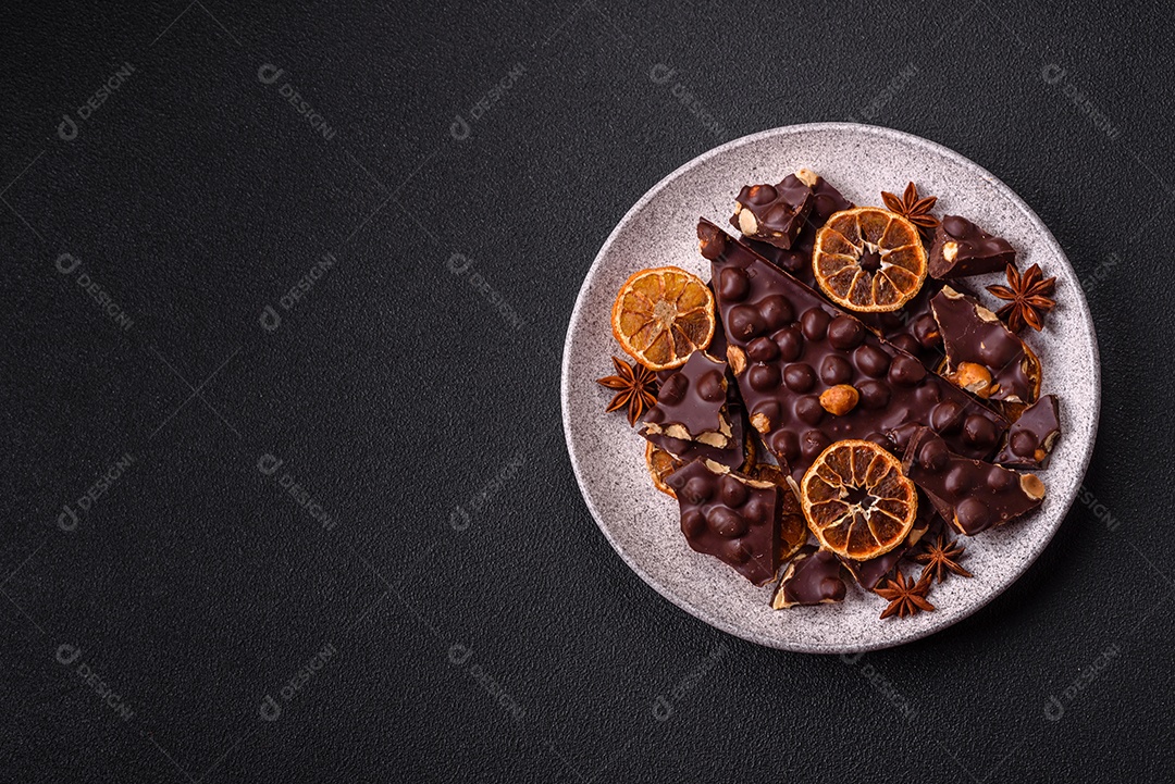 Chocolate amargo preto com avelãs inteiras, frutas cristalizadas e hortelã, quebrado em pedaços sobre fundo escuro de concreto