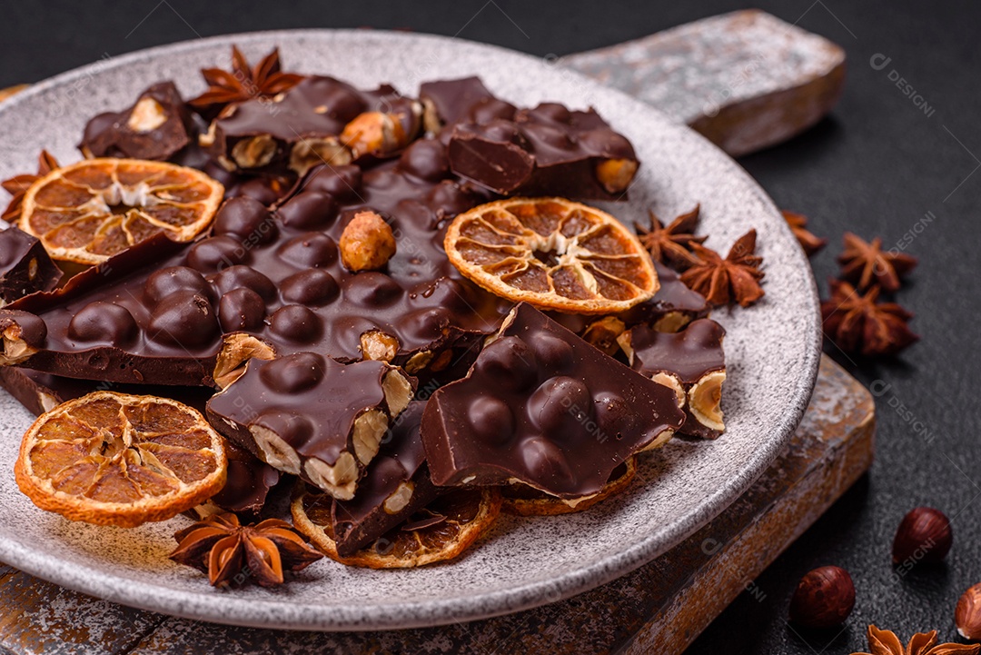 Chocolate amargo preto com avelãs inteiras, frutas cristalizadas e hortelã, quebrado em pedaços sobre fundo escuro de concreto