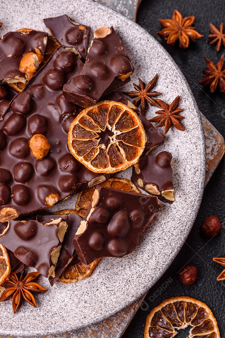 Chocolate amargo preto com avelãs inteiras, frutas cristalizadas e hortelã, quebrado em pedaços sobre fundo escuro de concreto