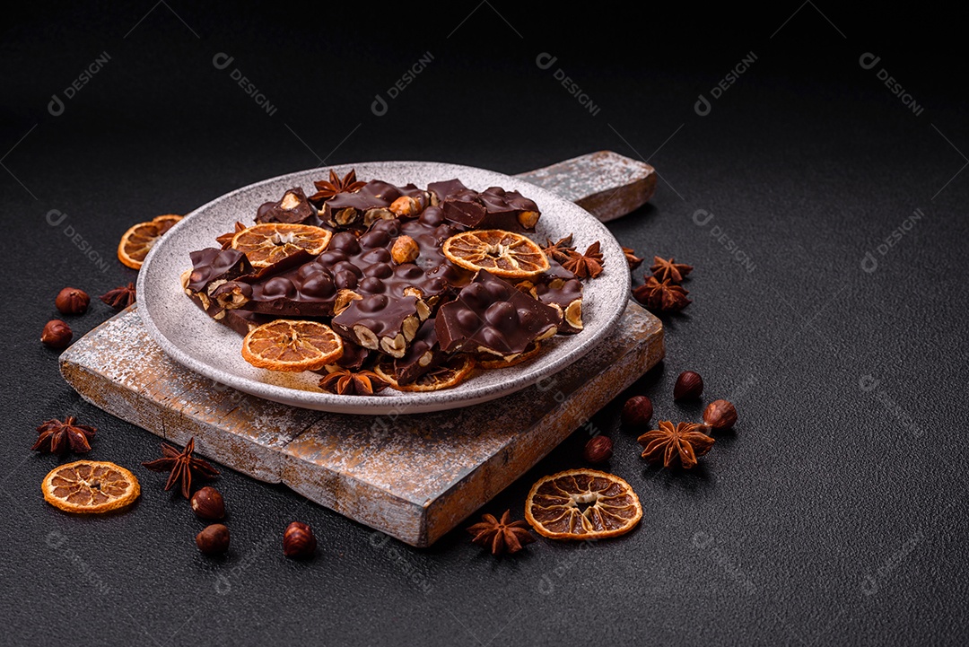 Chocolate amargo preto com avelãs inteiras, frutas cristalizadas e hortelã, quebrado em pedaços sobre fundo escuro de concreto