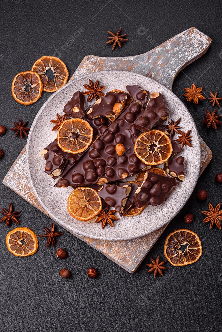 Chocolate amargo preto com avelãs inteiras, frutas cristalizadas e hortelã, quebrado em pedaços sobre fundo escuro de concreto