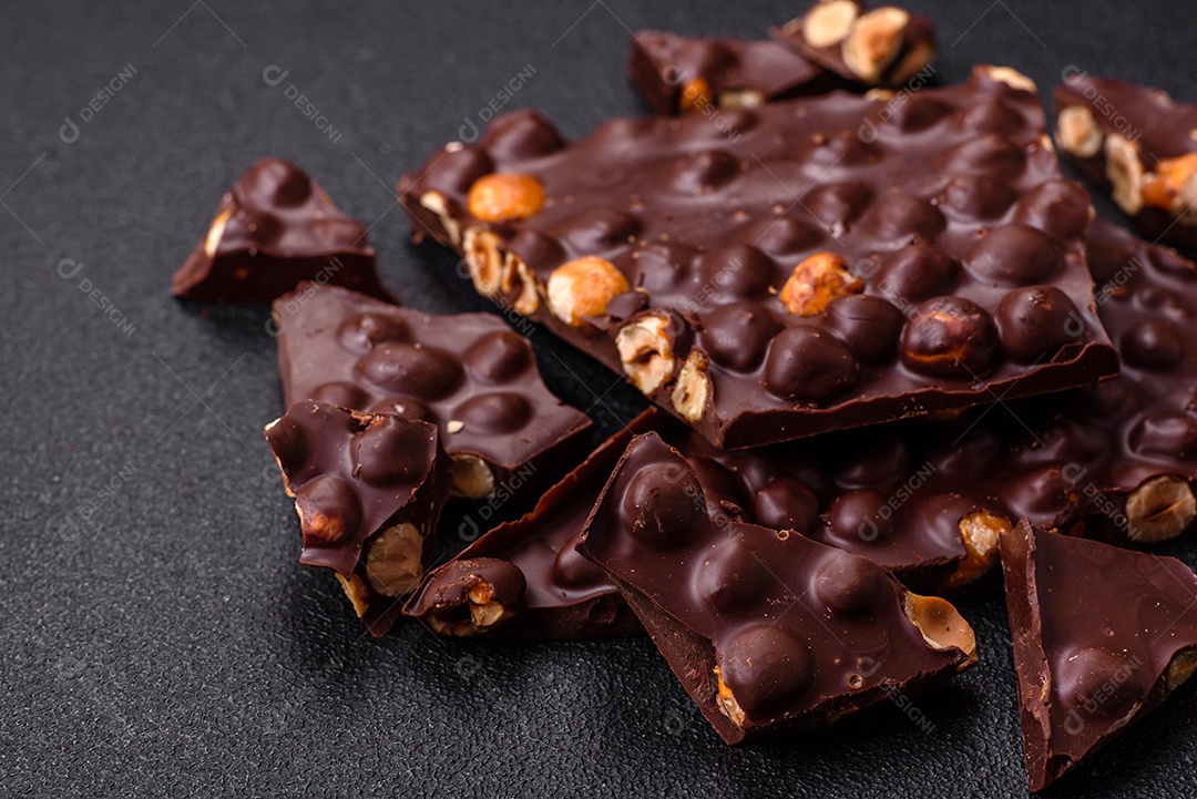 Chocolate amargo preto com avelãs inteiras, frutas cristalizadas e hortelã, quebrado em pedaços sobre fundo escuro de concreto