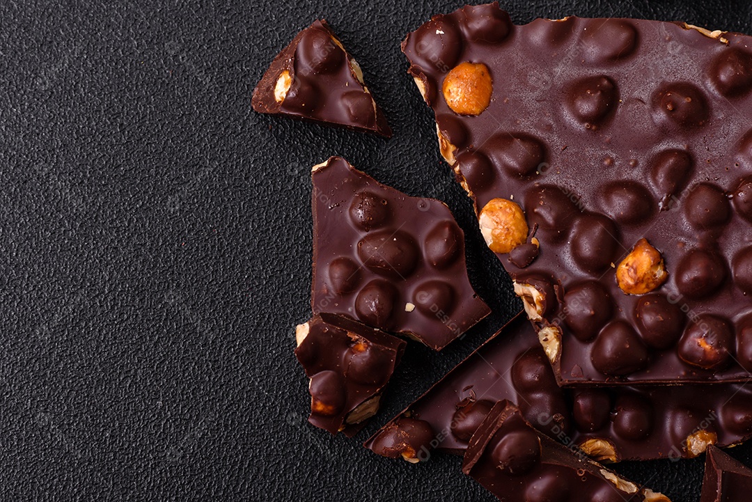 Chocolate amargo preto com avelãs inteiras, frutas cristalizadas e hortelã, quebrado em pedaços sobre fundo escuro de concreto