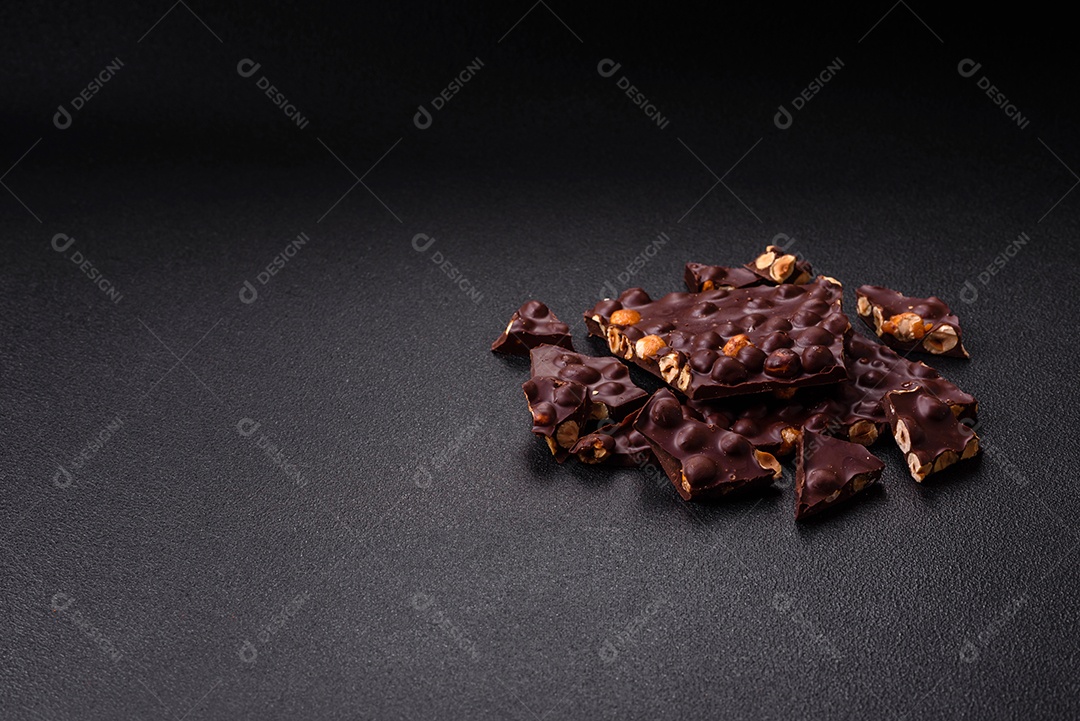 Chocolate amargo preto com avelãs inteiras, frutas cristalizadas e hortelã, quebrado em pedaços sobre fundo escuro de concreto